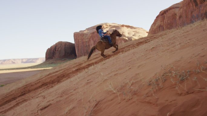 日落时分，在亚利桑那州/犹他州的纪念碑谷沙漠中，一个十几岁的美国原住民女孩（纳瓦霍人）骑着马爬上陡峭