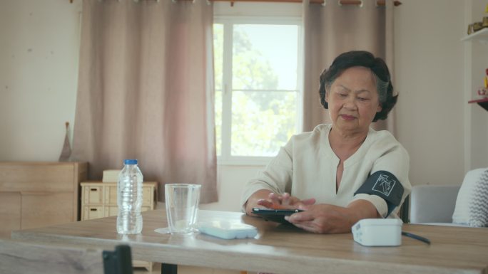 这位资深女士正在家里用数字血压计检查血压和心率，并将检测结果记录在平板电脑上。