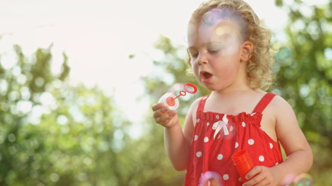 穿着红色连衣裙的斯洛莫蹒跚学步的小女孩在阳光明媚的果园里吹泡泡