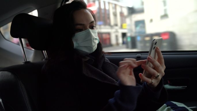 在旅途中，戴着口罩的女士正在汽车后座使用智能手机。