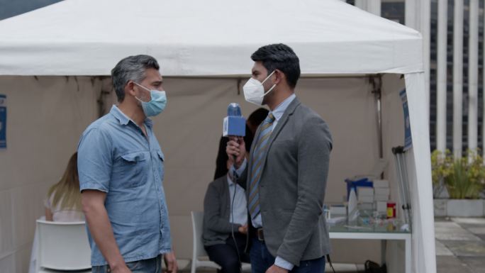 拉丁美洲男性新闻记者在新冠肺炎疫苗接种站采访一名等待接种疫苗的患者