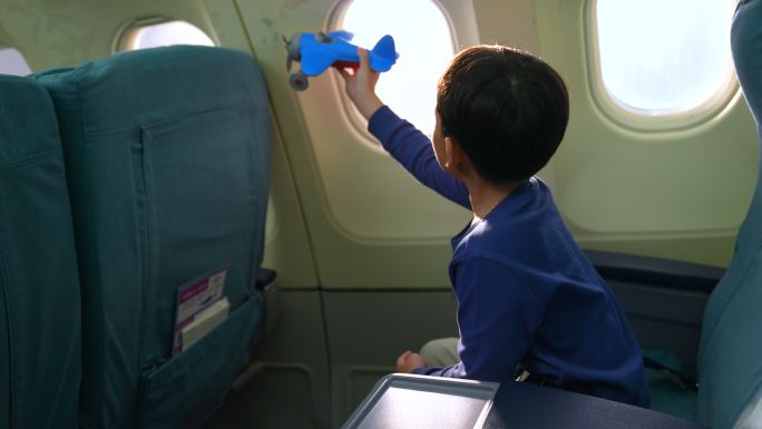一个男孩坐在商用飞机旁的窗户上。他手里拿着一架玩具飞机，梦想着坐飞机旅行或成为一名飞行员。
