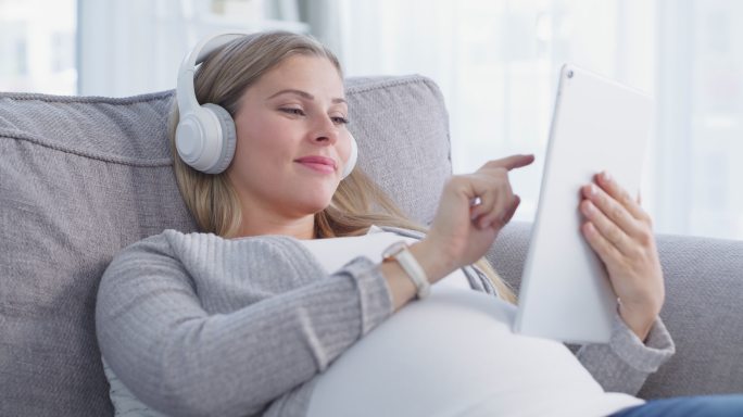 一个放松的怀孕年轻女子在家沙发上使用数字平板电脑和耳机的4k视频片段