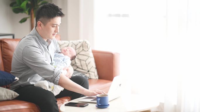 一名亚裔中国男子背着坐在沙发上的男婴在客厅里工作