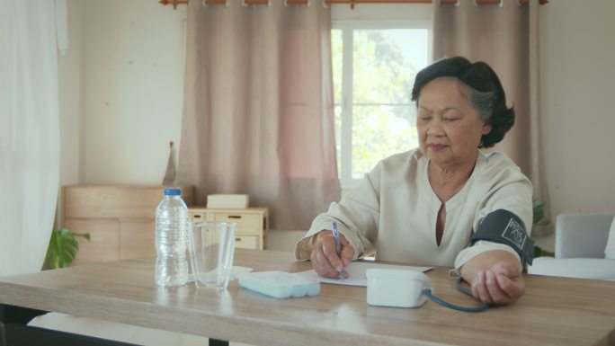 这位年长的女士正在家里用数字血压计检查血压和心率，并将测试结果记录在纸上。
