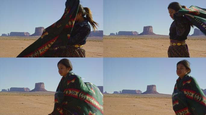 在一个晴朗的日子里，一个十几岁的美国原住民女孩在纪念碑谷沙漠中用传统的纳瓦霍人毯子裹着肩膀，远处有巨