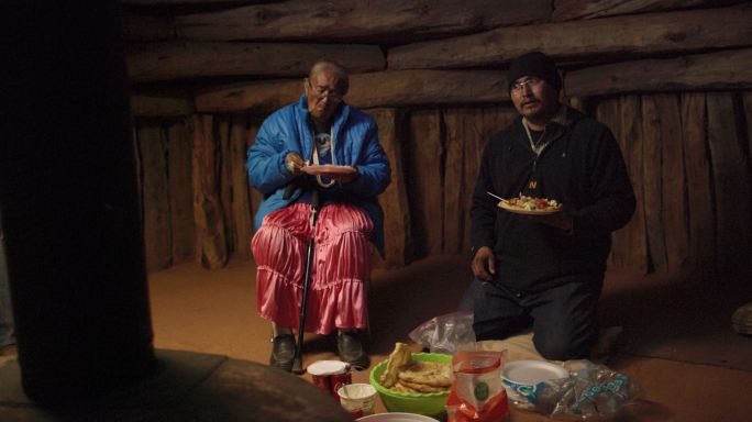 一名40多岁的美国原住民男子（纳瓦霍人）跪在霍根（纳瓦霍人小屋）的地板上，一边吃着食物，一边与其他人