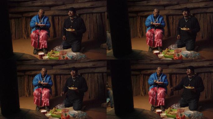 一名40多岁的美国原住民男子（纳瓦霍人）跪在霍根（纳瓦霍人小屋）的地板上，一边吃着食物，一边与其他人
