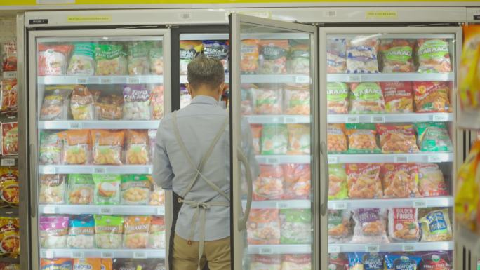 戴着口罩的亚裔中国高级男子在超市冷藏区用数字平板电脑清点冷冻食品