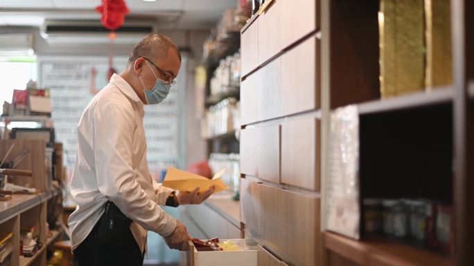 亚裔中国男药剂师在中药店从托盘中挑选和包装中药