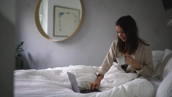 年轻女子一边在床上喝咖啡一边在笔记本电脑上浏览