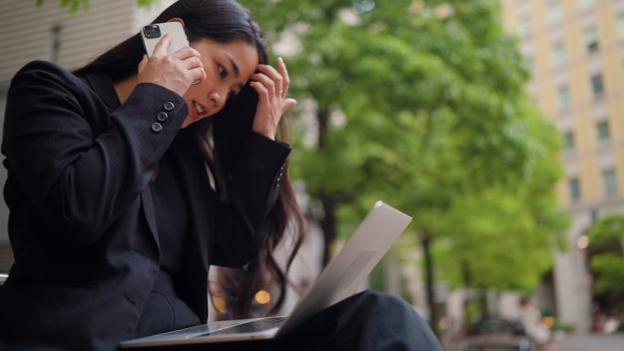在城市里，女商人坐在长凳上，一边用笔记本电脑一边打电话