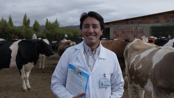 拉丁美洲男性兽医在一个畜牧场与奶牛一起工作，微笑着看着摄像机