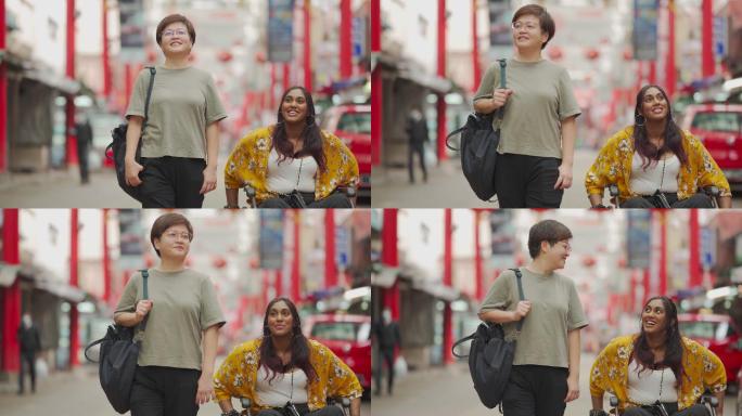 在城市街上，一名亚裔华裔中年女子和她的印度残疾女性朋友坐在轮椅上并肩行走