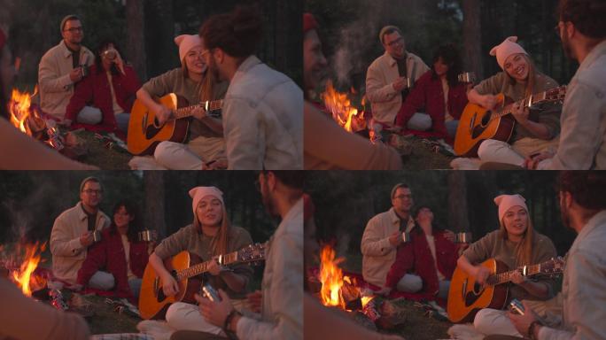 在篝火旁露营时，一个快乐的女人给她的朋友们弹吉他