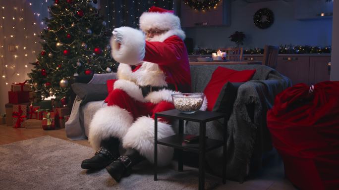圣诞老人坐在装饰精美的客厅里，坐在灰色沙发上看电视，吃爆米花。圣诞老人挥舞着拳头，对电影的情节做出情