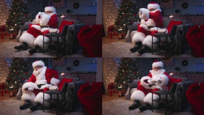 圣诞老人坐在装饰精美的客厅里，坐在灰色沙发上看电视，吃爆米花。圣诞老人挥舞着拳头，对电影的情节做出情