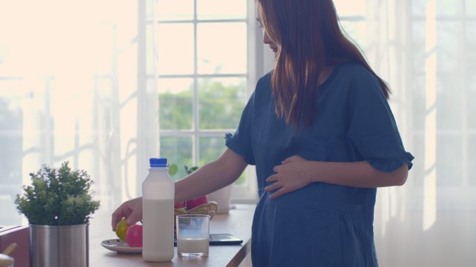 一位孕妇正在喝牛奶，轻轻地触摸她的腹部，怀孕/分娩