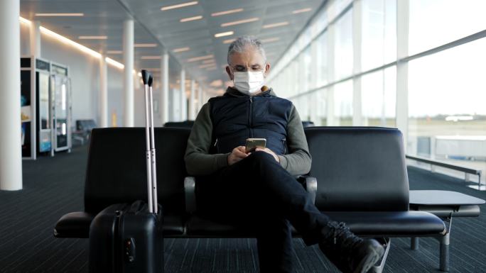 戴着防护面罩的中年男子坐在机场候机大厅里，提着行李箱，手里拿着手机。