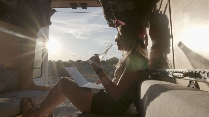 科罗拉多州弗鲁塔附近，一位金发碧眼的千禧一代年轻女子开着定制露营车，用笔记本电脑从瓶子里喝水