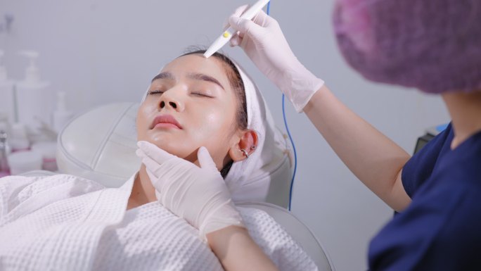 亚洲年轻美丽的女性，穿着浴衣，头戴毛巾头巾，躺在医生面前，在额头皮肤上做皮肤针疗法。亚洲年轻美丽的女