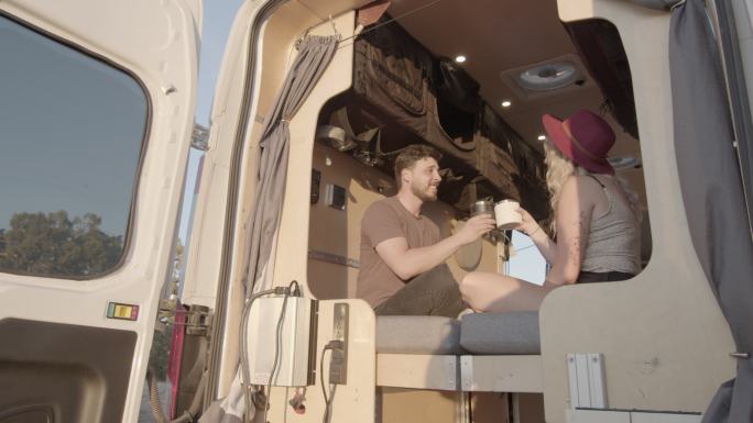 视频剪辑显示，一对年轻夫妇坐在他们的面包车里，开着后门看日落，一边聊天，一边喝着一杯咖啡