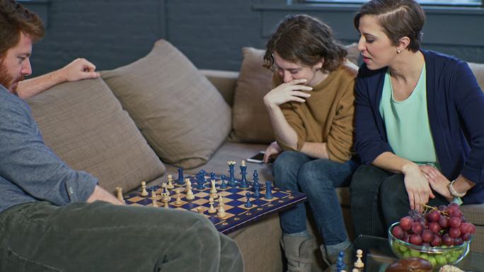 成熟的男人在客厅里和十几岁的女孩下棋，而她的姐姐给了她一些关于这项运动的建议