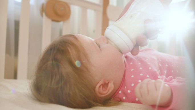在一个阳光明媚的早晨，穿着粉色紧身衣的可爱快乐女婴在室内喝着婴儿奶瓶里的牛奶