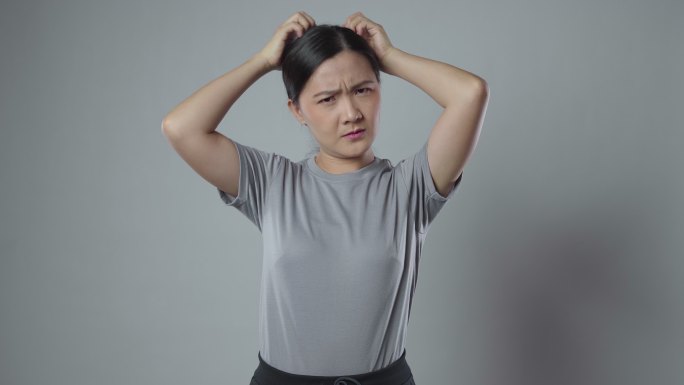 一名亚洲女子搔着头，孤零零地站在灰色背景上。