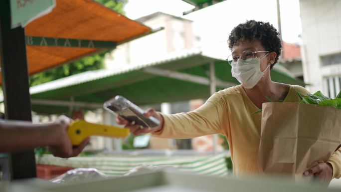 在农贸市场使用非接触式支付方式支付食品杂货的年轻女性——戴着防护口罩