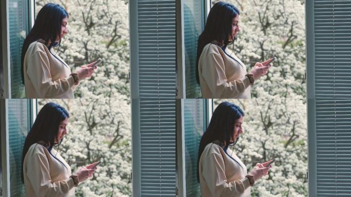 春天，年轻女子在窗前用手机放松。背景是盛开的果树。