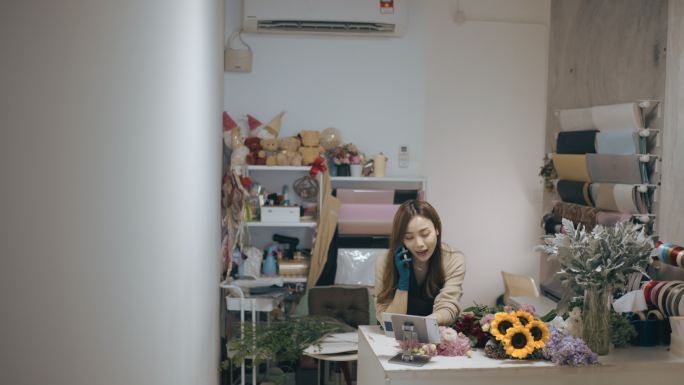 亚裔中国女性花商，小型企业花店老板，在工作场所使用数字平板电脑时用智能手机交谈。检查库存、接受客户订