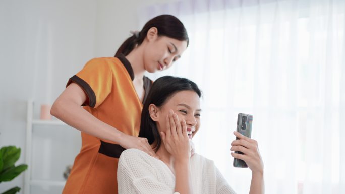 穿着浴袍的亚洲美女坐在水疗沙龙里接受年轻专业按摩师的泰式背部和颈部按摩，用视频电话谈论按摩。美容治疗