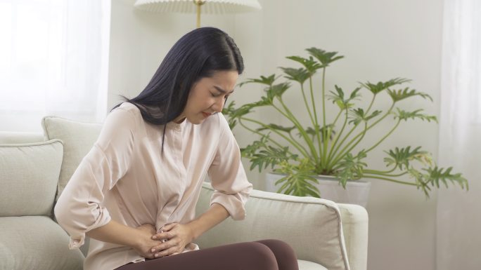 亚洲女性因胃痛坐在沙发上。她在家里胃痛得厉害。