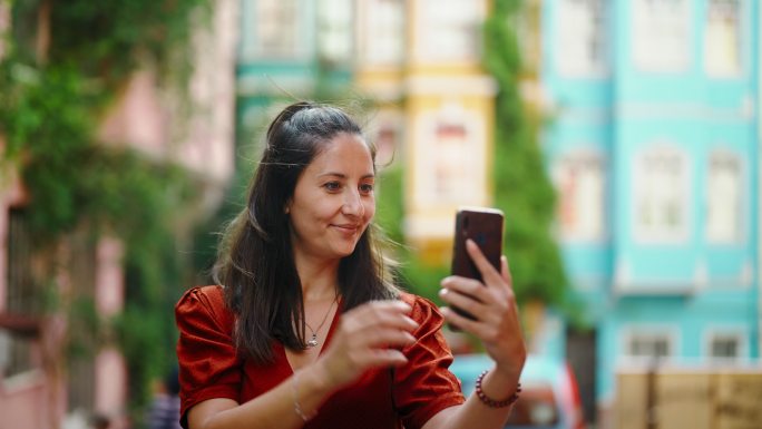 穿着红色长裙的美女在城市的彩色建筑前使用智能手机