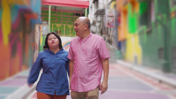 亚裔马来退休父亲和他的唐氏综合症女儿在周末的休闲活动中手牵着手走在城市街道上