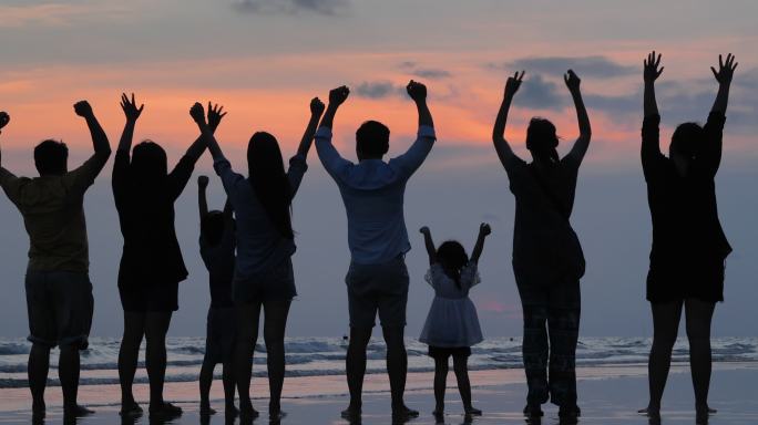 后视图，在剪影中，一个由八个人组成的多代人家庭在仰望风景的同时向空中举起双臂，张开双臂在日落时分的海
