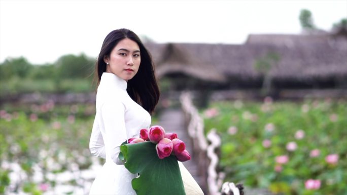 4K慢镜头FHD拍摄了一幅美丽的越南女子手持粉色莲花，在越南大莲花湖木桥上微笑的画面，这是一个亚洲或
