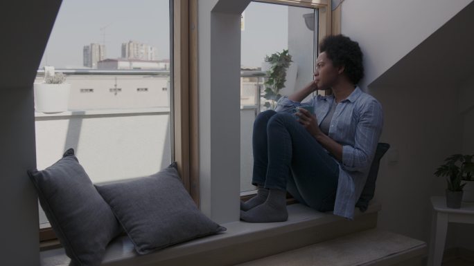 妇女坐在公寓的窗台上，端着一杯茶沉思着。