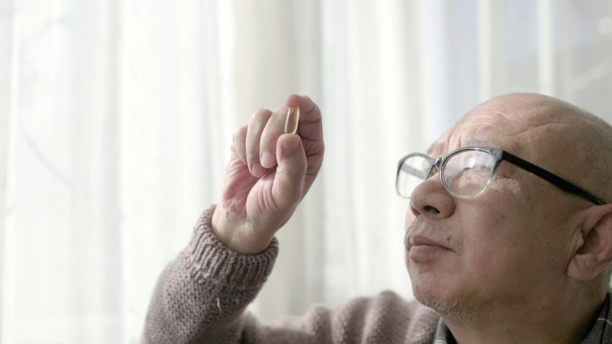 一位坐在窗边的中国老人手里拿着一个胶囊，戴着阅读眼镜观看