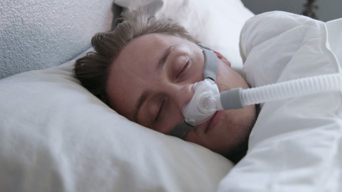 一名患有睡眠呼吸暂停症的年轻男子的特写镜头，他戴着CPAP面罩躺在床上侧卧