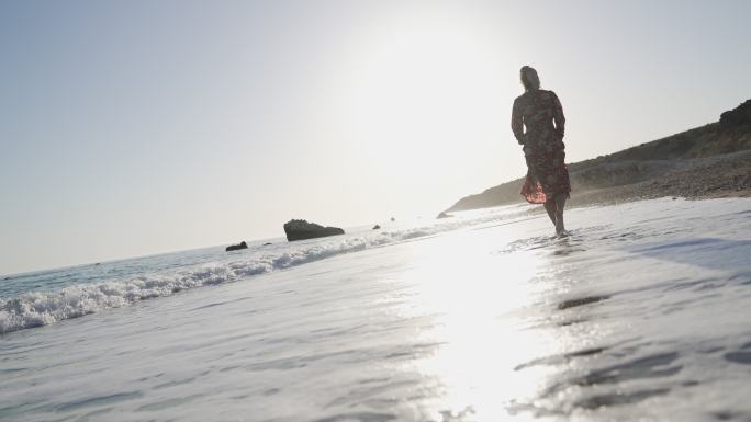 宁静的赤脚女人穿着裙子，日落时在海滩上散步，海浪拍打着她的脚，她享受着每一刻