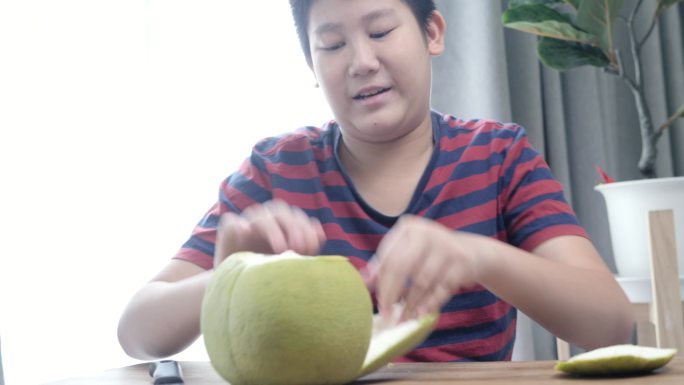 快乐的亚洲男孩坐在家里窗户附近的地板上剥柚子，这是一种生活方式。