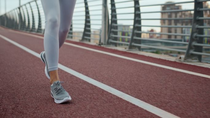 一段慢镜头视频，一名女性跑步者穿着白色紧身裤和灰色运动鞋在跑道上慢跑