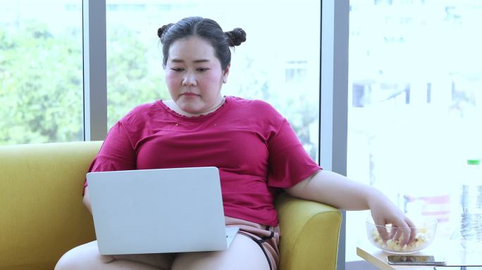 身材高大的女士使用笔记本电脑和平板电脑，亚洲女孩喜欢在家里的沙发上吃东西