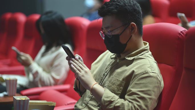 亚裔-华人混合年龄组的人坐在电影院里用智能手机等待电影上映时间