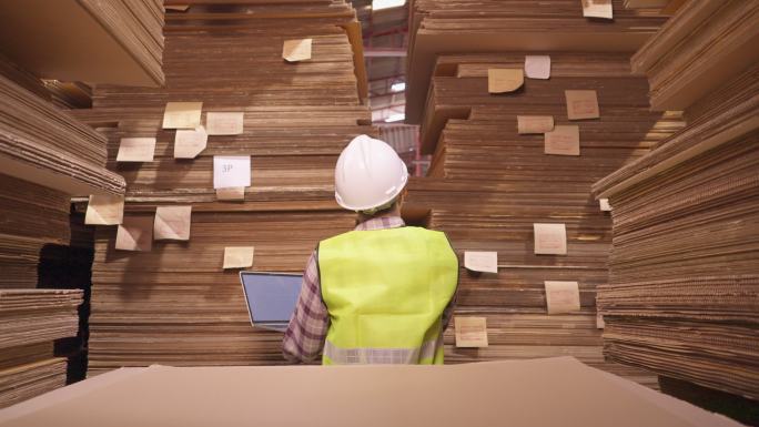 后视图：亚洲纸包装制造工人。使用手提电脑在安全背心和头盔包装制造的纸板堆中间进行检查表和盘点