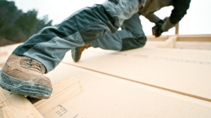 一名工人的脚站在屋顶板条上，一边用螺丝钉穿过屋顶上的木梁