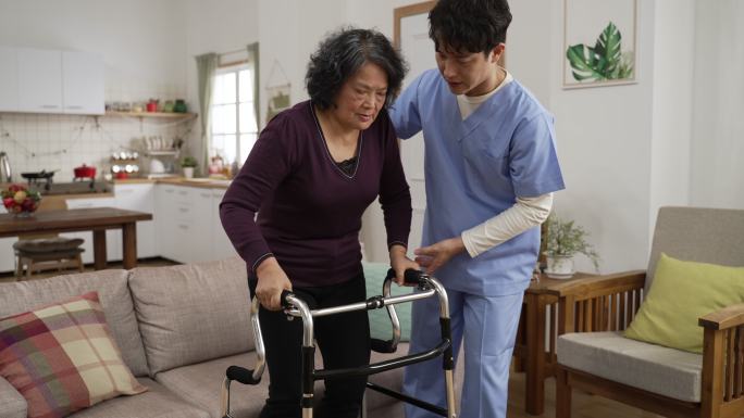 亚洲男性个人护理助理帮助虚弱的成熟女性在家里的客厅里与助行器一起行走。在家照顾受伤老人的理念