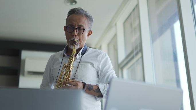 lifehack亚洲活跃的高级男子艺术家演奏萨克斯管，并在客厅用笔记本电脑向学生展示
